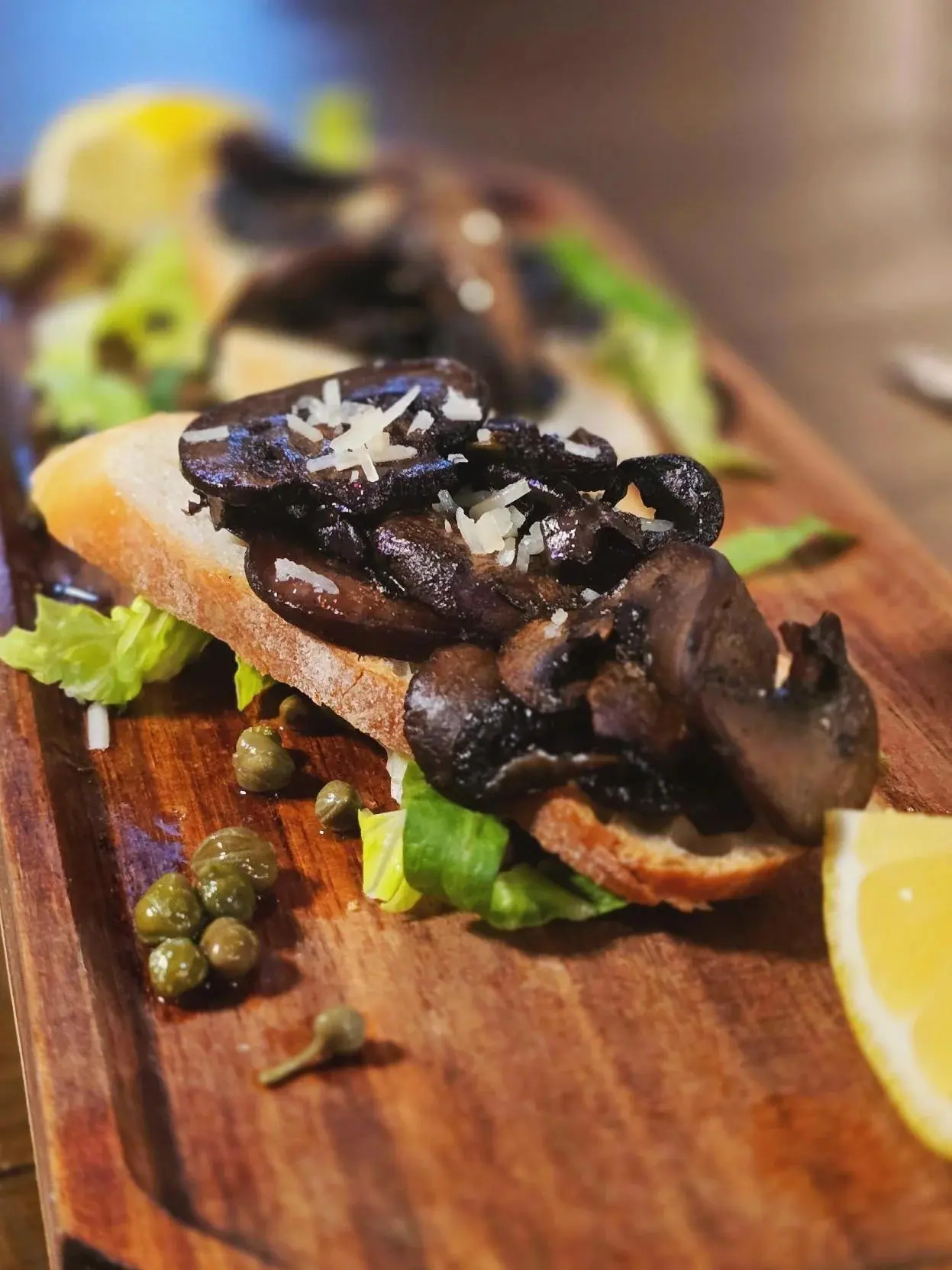 Close-up of roasted mushroom crostini with shaved cheese, capers and greens on a walnut board