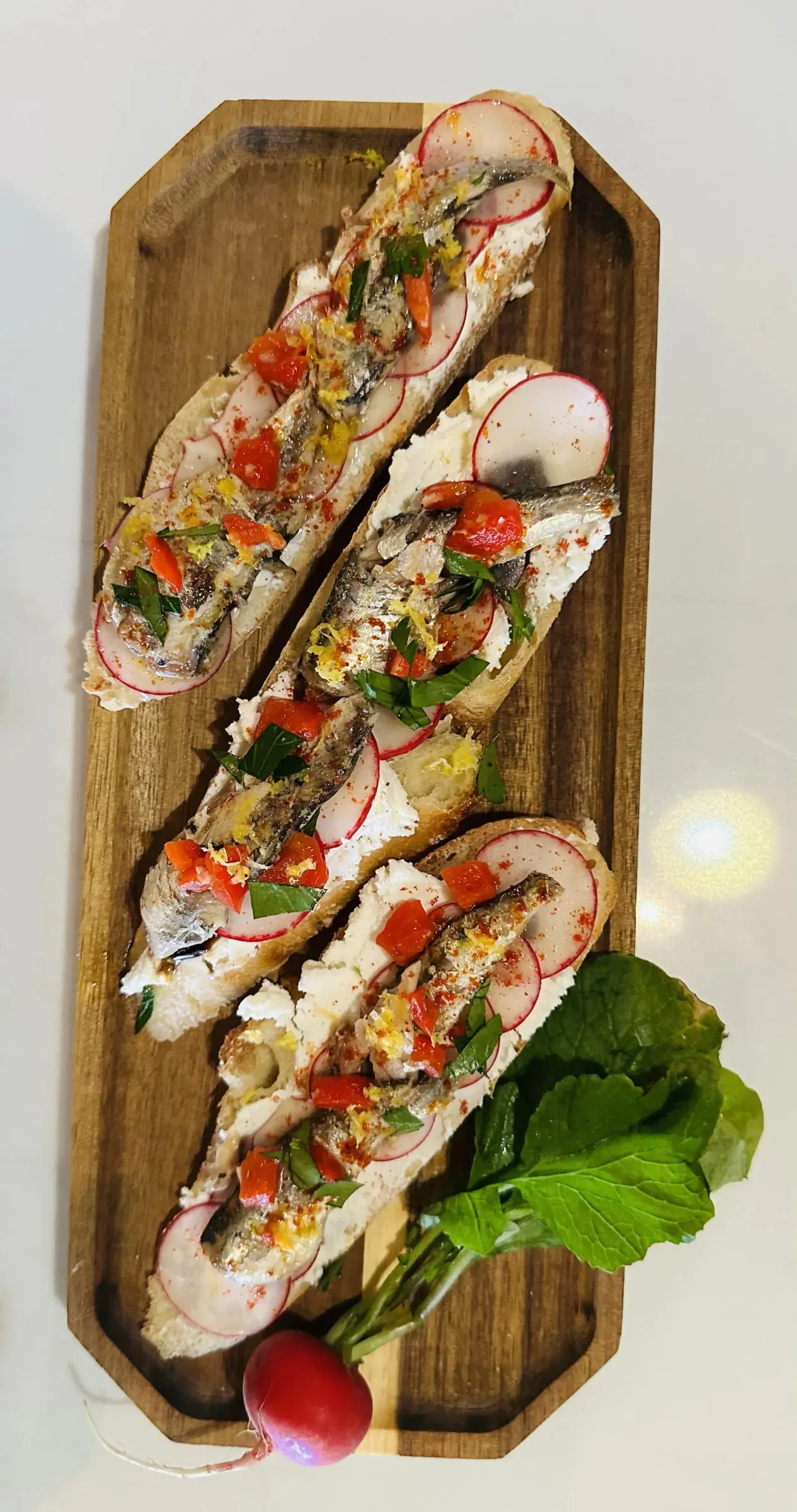 Open-faced sardine toasts with whipped cheese, shaved radish, peppers and lemon zest on a wooden board